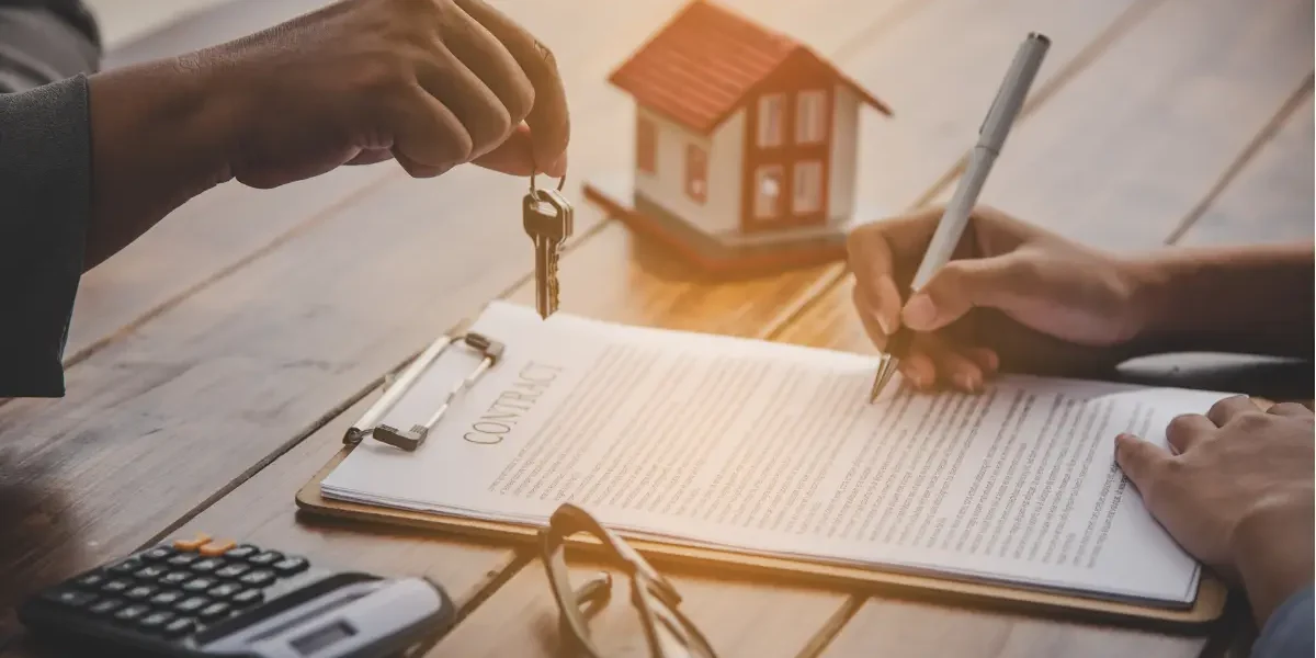 A person signing a real estate contract on a wooden table while an agent hands over a set of house keys, with a small model house in the background.
